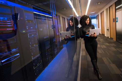 Photo of a woman with a laptop in a corridor leaning against server racks.